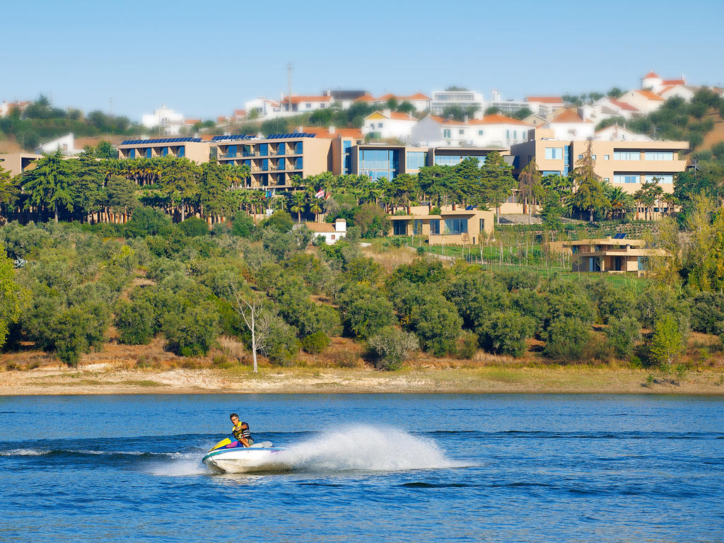 Ap Lago Montargil - Conference & Spa Luaran gambar The photo depicts a person riding a jet ski on a body of water. In the background, there are modern buildings and lush greenery, suggesting a scenic landscape, likely near a resort or upscale residential area. The clear blue sky and calm water enhanc