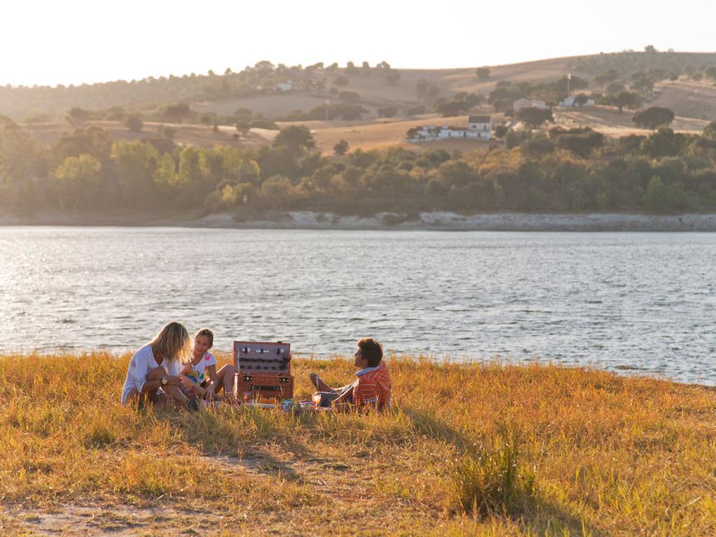 Ap Lago Montargil - Conference & Spa Luaran gambar The photo depicts a serene lakeside scene during sunset. Three people are gathered on a grassy area near the water, enjoying a picnic. They are seated around a picnic basket, with the shimmering lake in the background and hills in the distance. The w