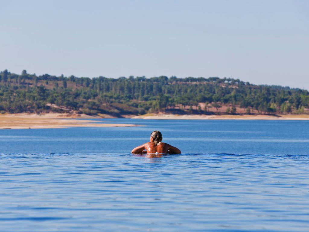 Ap Lago Montargil - Conference & Spa Luaran gambar The photo shows a person swimming in a calm body of water, likely a lake or a river. The swimmer is seen from behind and appears to be enjoying the water on a sunny day. In the background, there's a scenic landscape featuring green trees and a sandy 