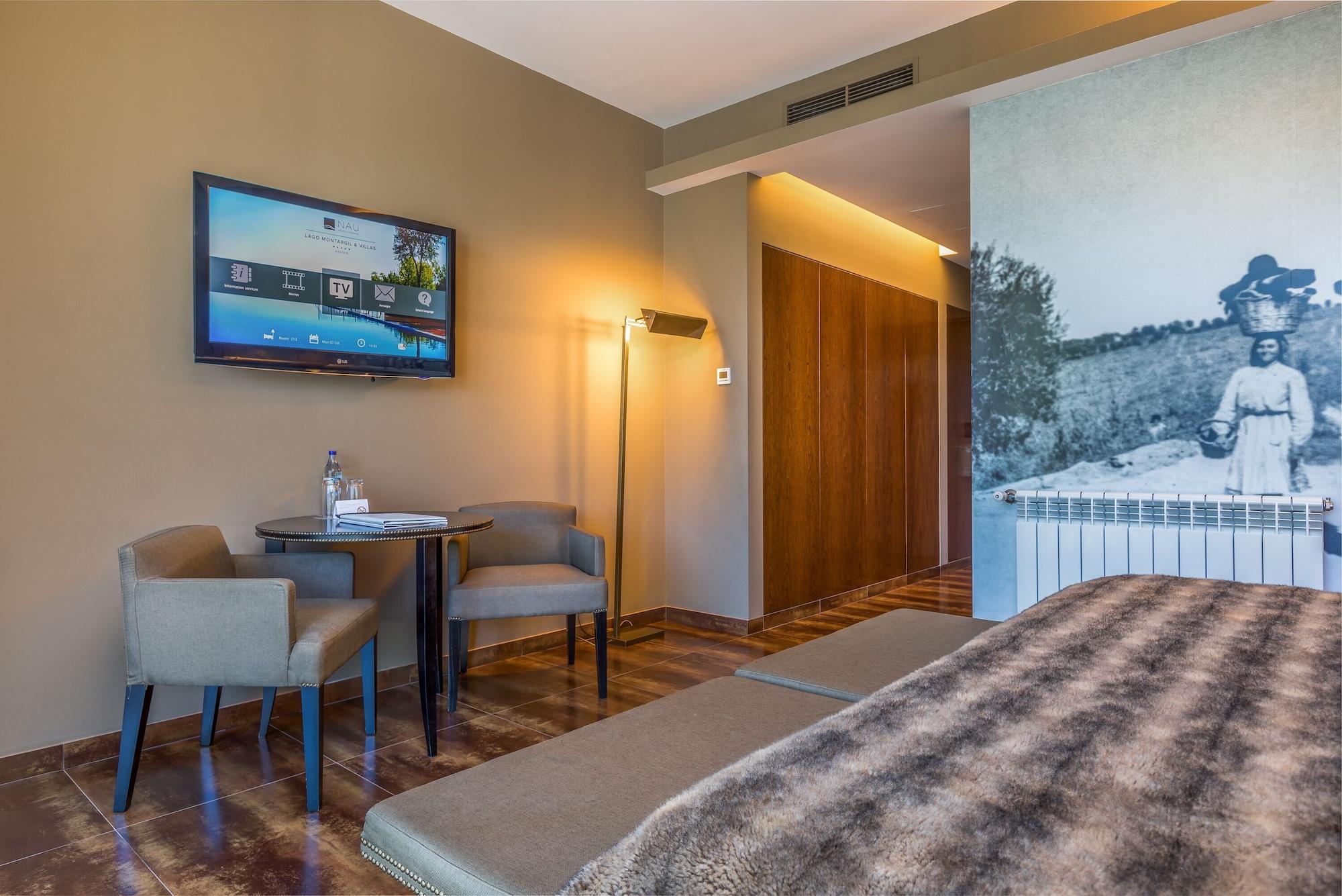 Ap Lago Montargil - Conference & Spa Luaran gambar The photo shows an interior of a room, likely a hotel or a modern living space. In the foreground, there is a bed with a textured brown blanket. To one side, there are two comfortable chairs and a small table, possibly with a bottle of water. A flat-