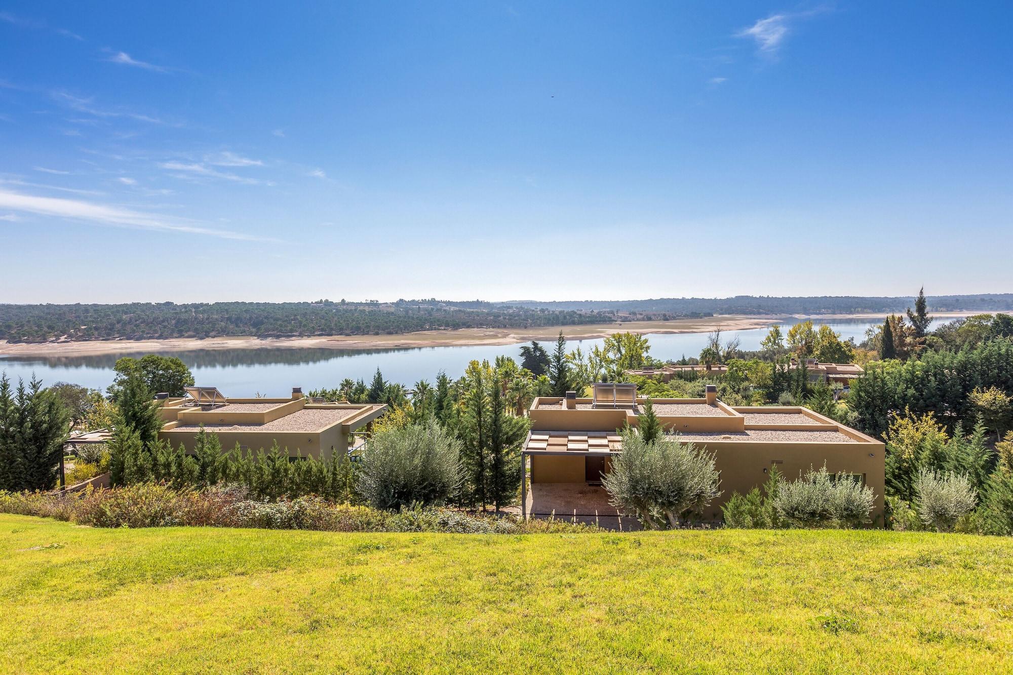 Ap Lago Montargil - Conference & Spa Luaran gambar The photo depicts a scenic landscape featuring a calm body of water, possibly a lake or river, surrounded by lush greenery and trees. In the foreground, there are several modern buildings with flat roofs, set against a vibrant blue sky. The overall s