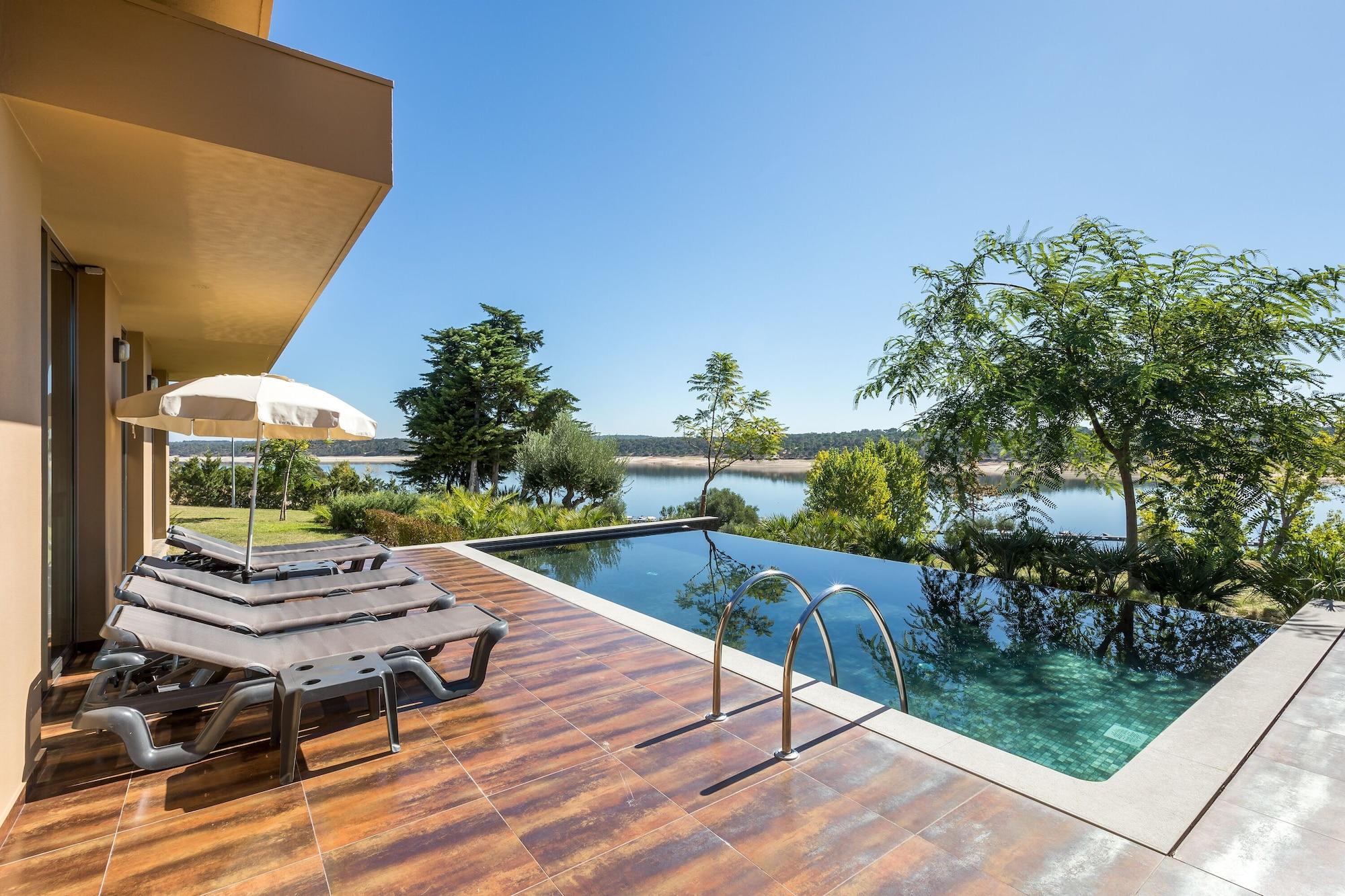 Ap Lago Montargil - Conference & Spa Luaran gambar The photo shows a luxurious outdoor setting by a pool. There are several lounge chairs arranged next to an infinity pool, which overlooks a scenic body of water and lush greenery. A parasol provides shade for relaxation. The sky is clear and blue, in