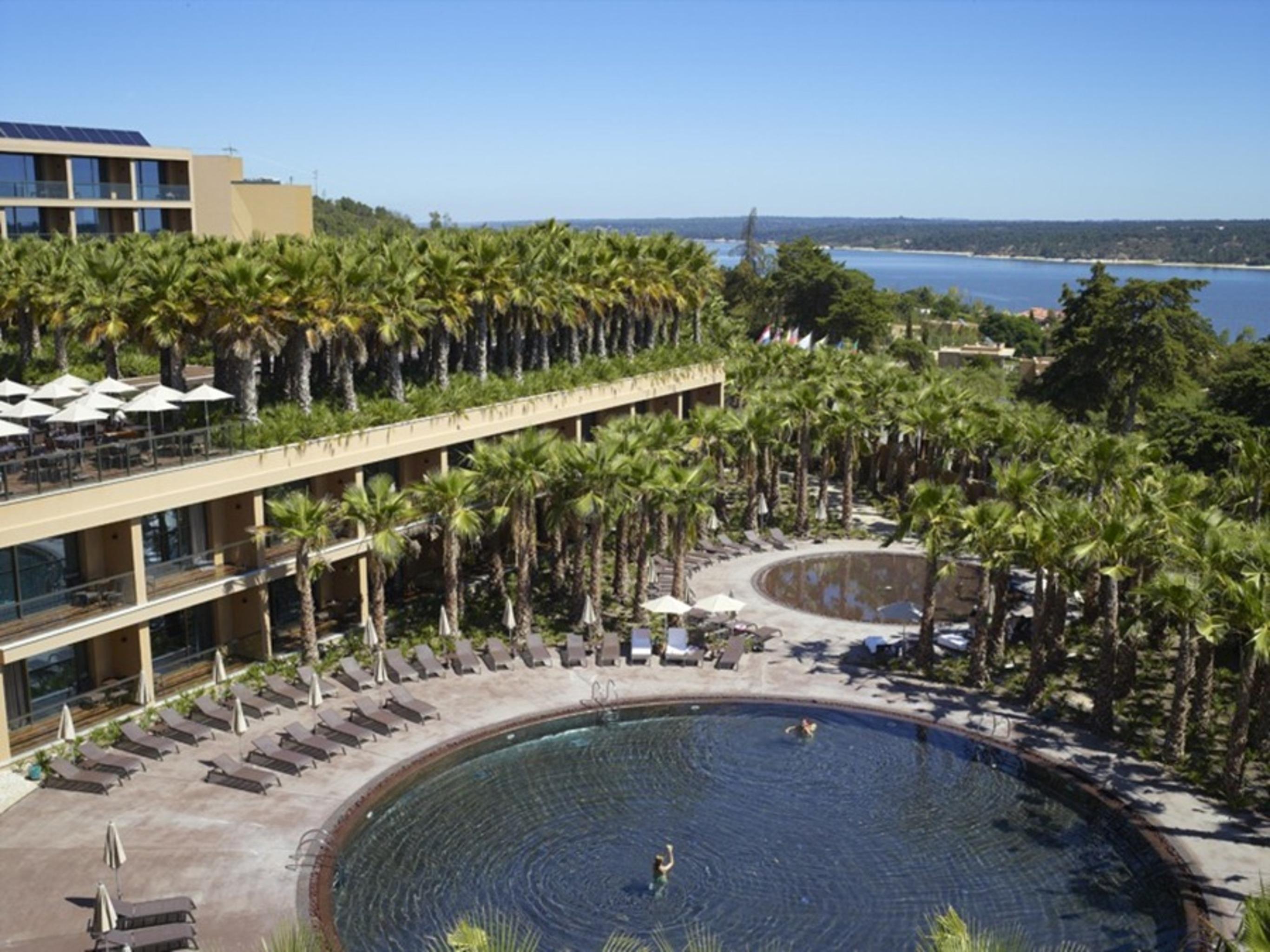 Ap Lago Montargil - Conference & Spa Luaran gambar The photo shows a scenic view of a resort area. In the foreground, there are circular pools surrounded by lounge chairs, with a few people enjoying the water. Palm trees line the edges, creating a tropical atmosphere. In the background, there is a mu