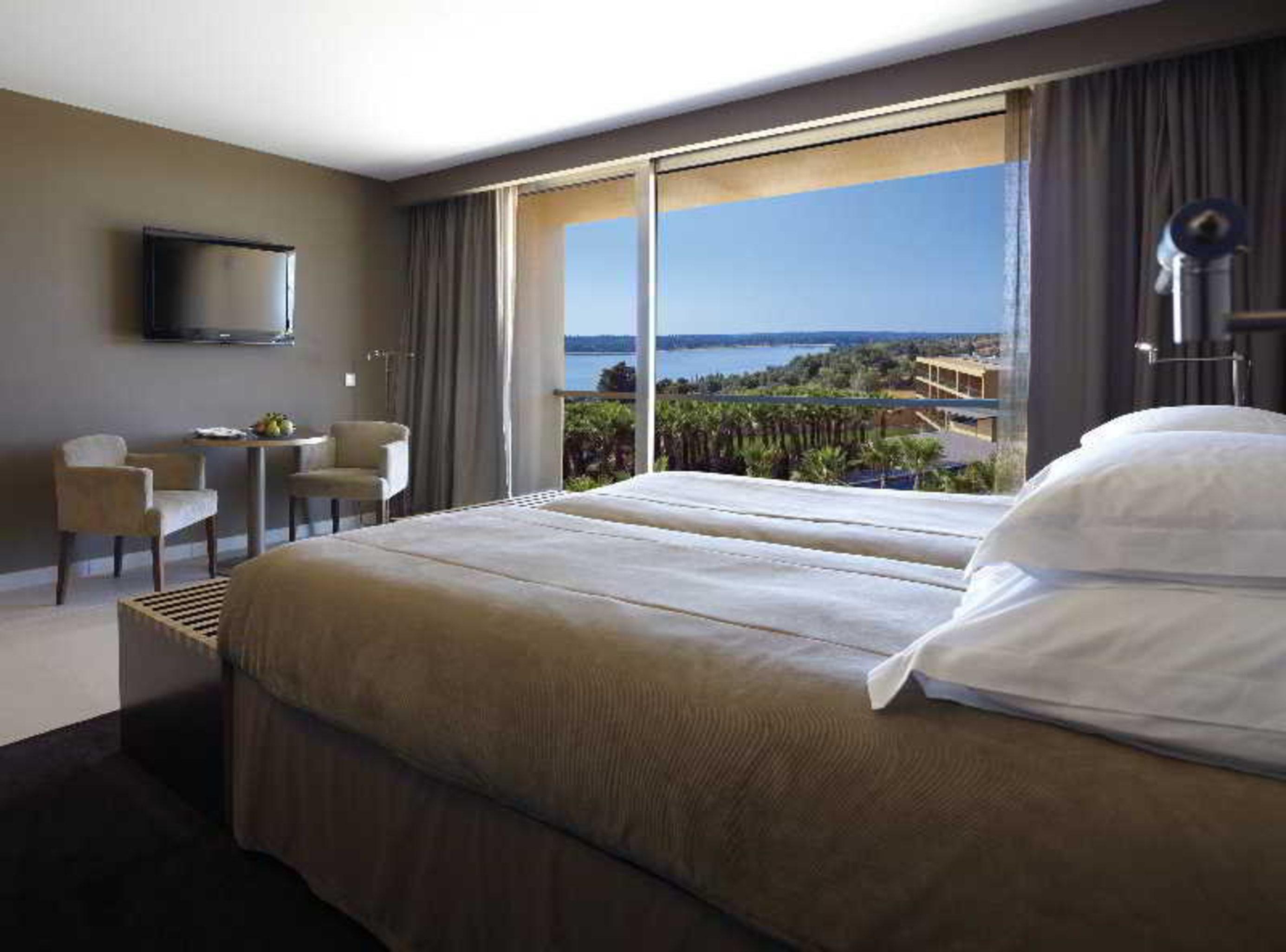 Ap Lago Montargil - Conference & Spa Luaran gambar The photo shows a modern hotel room with a large window offering a view of the sea and mountains. The room features a comfortable bed with white pillows and brown bedding. There is a small sitting area with two chairs and a table, as well as a wall-m
