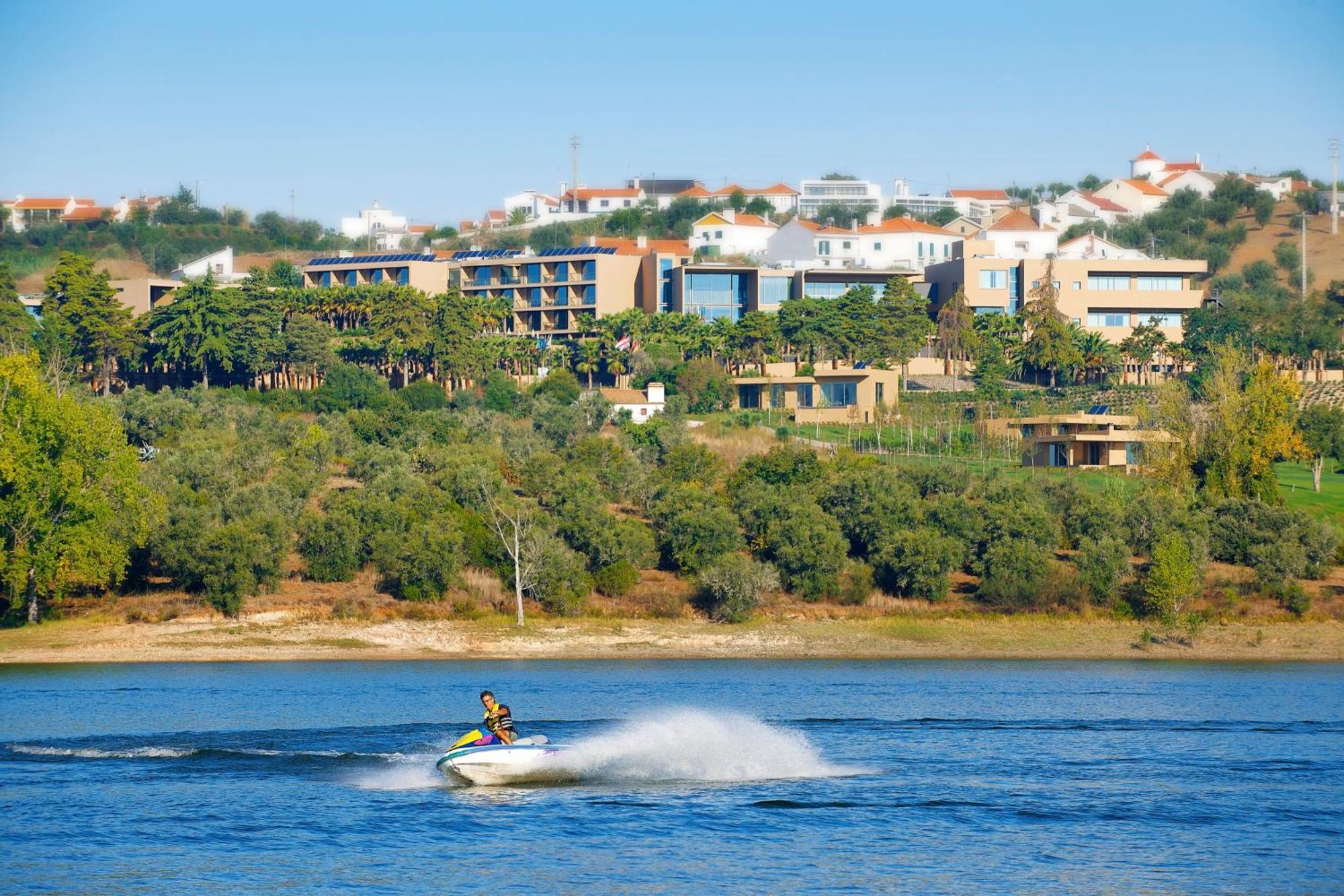 Ap Lago Montargil - Conference & Spa Luaran gambar The photo shows a person riding a jet ski on a body of water, creating a spray as they speed across the surface. In the background, there are modern buildings and a hillside with trees and greenery. The sky is clear and blue, suggesting a bright and 