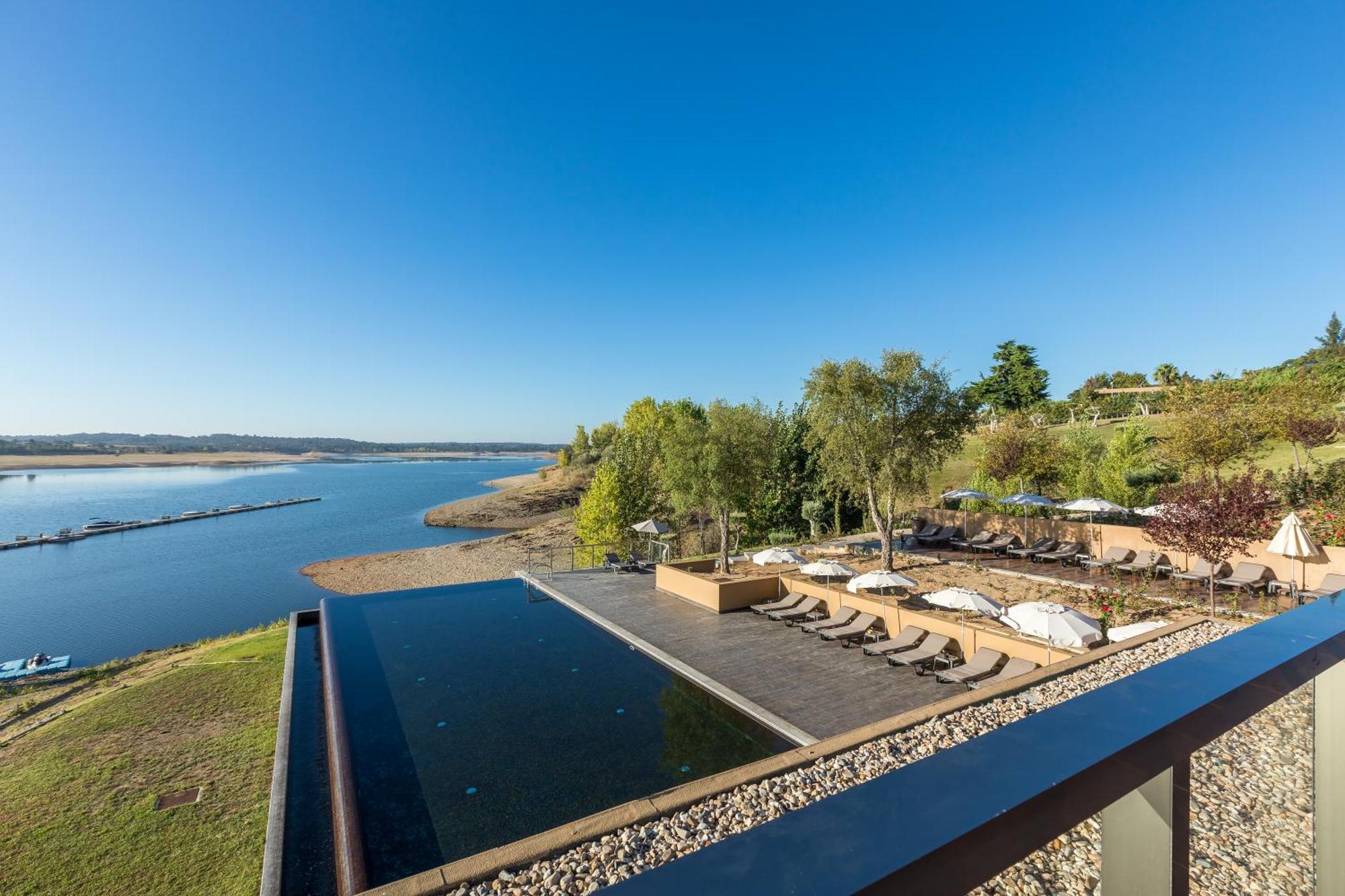 Ap Lago Montargil - Conference & Spa Luaran gambar The photograph depicts a scenic view of a tranquil water body, likely a lake or river, bordered by lush greenery. In the foreground, there is an infinity pool that blends seamlessly with the water beyond. Comfortable lounge chairs are arranged on a w