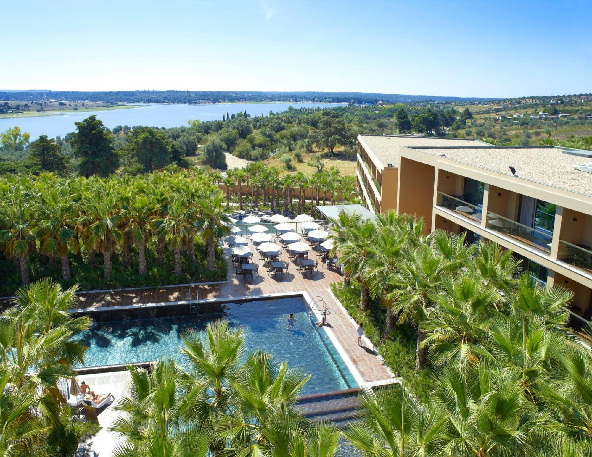 Ap Lago Montargil - Conference & Spa Luaran gambar The photo shows a scenic view of a modern hotel or resort. In the foreground, there is a swimming pool surrounded by palm trees, with lounge chairs positioned beside it. Sun umbrellas provide shade over the chairs. The background features a lush land